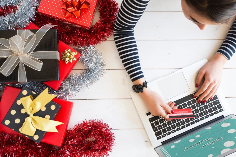 A woman using a laptop to shop online sitting next to wrapped presents.
